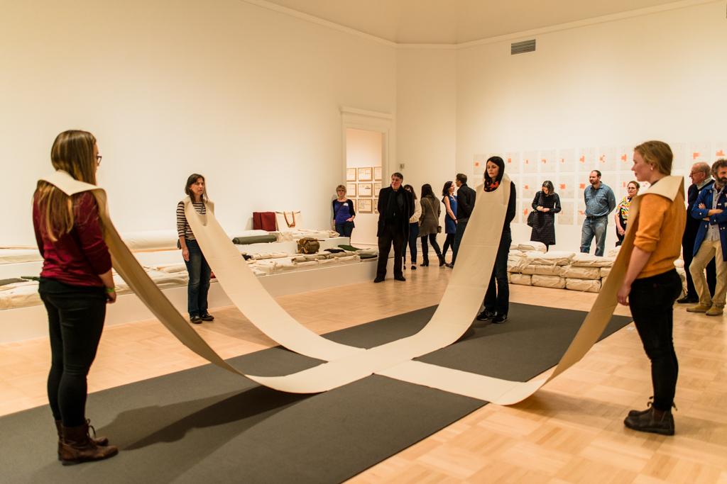 Four performers wearing a long cloth cross that connects them across a museum gallery, visitors watching in the background